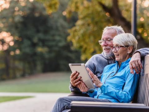 Koristekuva iäkkäämpi pariskunta istuu puistonpenkillä ja katsoo yhdessä tablet-tietokonetta.